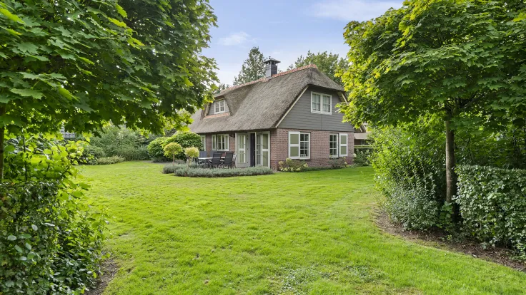 Rietgedekte villa met bakstenen gevel achter struiken en bomen op het gazon